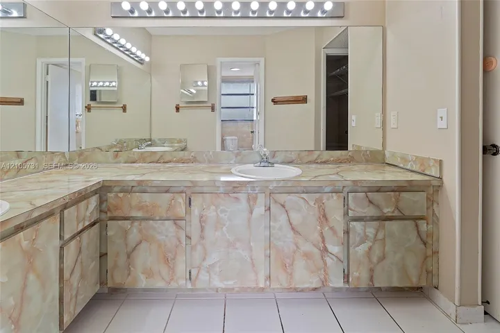 Double vanity featuring natural stone-patterned cabinetry and matching stone countertop with drop-in sinks, wall-to-wall mirror, and overhead multi-bulb lighting