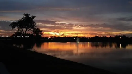 Waterfront property featuring a lake, active fountain, and mature tree silhouettes set against a sunset backdrop