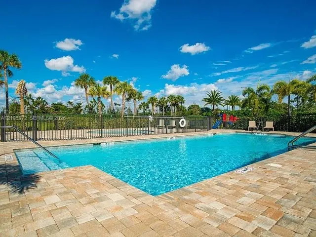 In-ground swimming pool featuring a brick paver deck, built-in entry steps, metal perimeter fencing, and mature palm trees