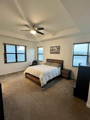 Tray ceiling with a central fan, multiple windows with neutral roller shades, and wall-to-wall carpeting