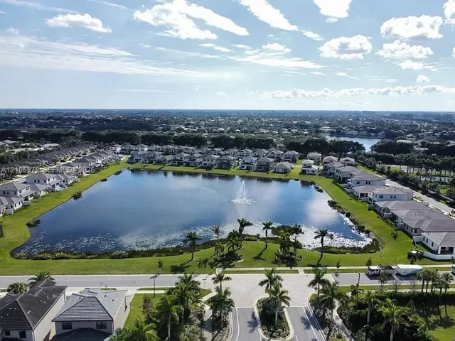 Aerial perspective of a master-planned community featuring a central lake with a fountain, paved street network, manicured landscaping, and mature palm trees