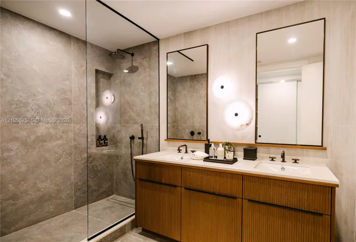 Double vanity with wood-finish cabinetry, stone-surface countertop, walk-in shower with large-format tile surround, frameless glass enclosure, and matte black wall-mounted hardware. Images are for finish purposes only. Please refer to the floor plan for the layout.