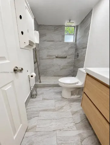 Bathroom featuring a walk-in shower with stone-pattern tile, a light-toned wood-finish vanity with a white countertop, and large-format porcelain floor tiling