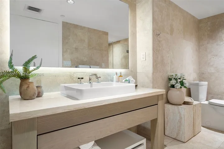 Bathroom featuring a stone-topped wood vanity, rectangular vessel sink, wall-spanning mirror with integrated under-lighting, and floor-to-ceiling stone tile wall finishes