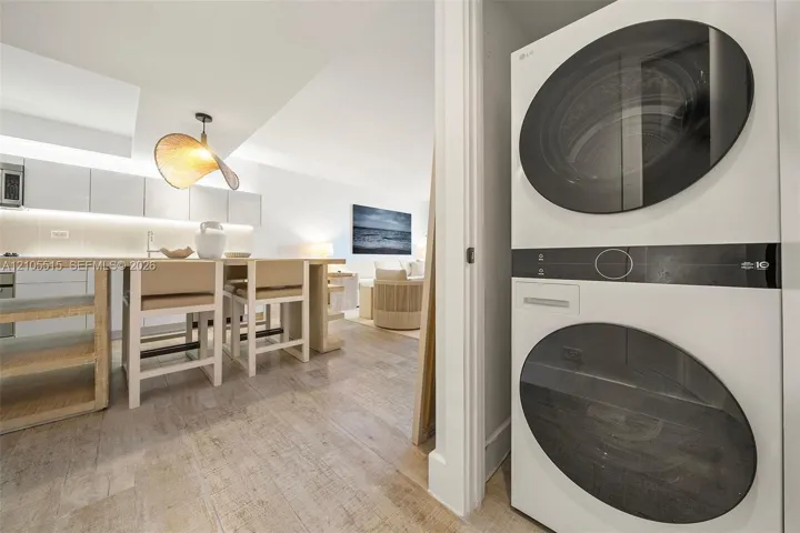 Stacked white laundry unit with round glass-front doors, featuring integrated kitchen cabinetry, wood-finish flooring, and a contemporary pendant lighting fixture