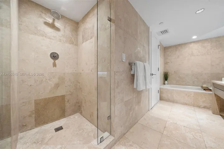 Walk-in shower featuring glass enclosure, rain-style showerhead, and natural stone tile surround, complemented by a stone-tiled soaking tub and recessed lighting