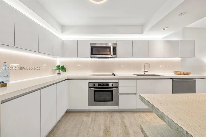 Kitchen featuring white flat-panel cabinetry, light-toned solid surface countertops, stainless steel appliances, integrated under-cabinet lighting, and wood-finish flooring