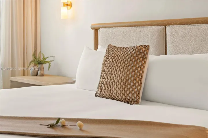 Custom wood-framed headboard with textured upholstery, light-toned wood-finish side table, and wall-mounted brass light fixture with frosted glass