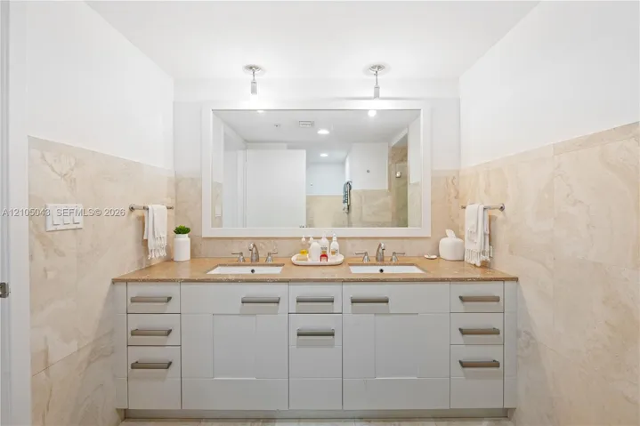 Double vanity featuring cabinetry with brushed metal hardware, dual under-mount sinks, integrated stone-finish countertop, and large wall-mounted mirror with integrated lighting