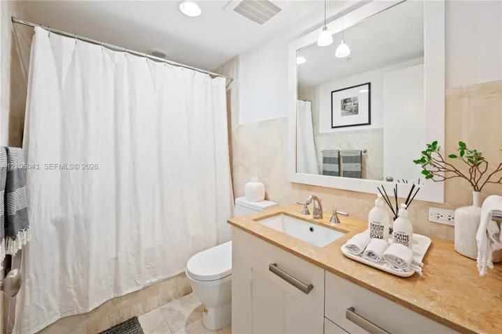 Bathroom featuring a white vanity cabinet with metallic hardware, light-toned stone countertop, rectangular undermount sink, framed mirror with integrated pendant lighting, and full-height stone wall tile
