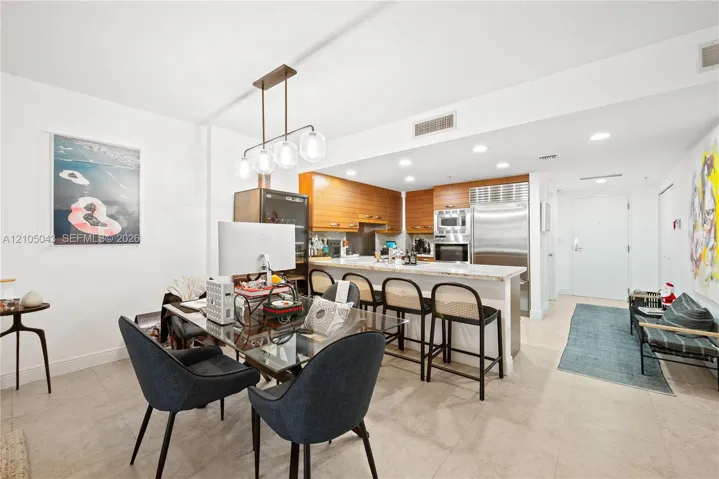 Kitchen featuring wood-toned cabinetry, stone countertops, stainless steel appliances, a multi-bulb linear pendant light, and neutral tile flooring