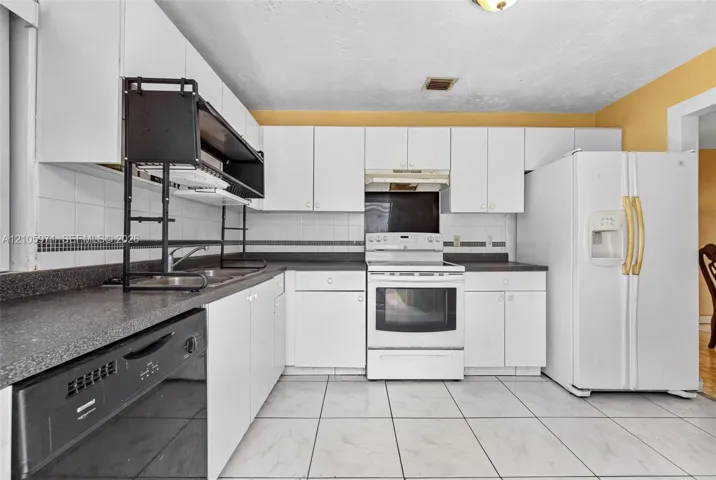 Kitchen featuring white cabinetry, dark countertops with tiled backsplash, white appliances, and large-format tile flooring