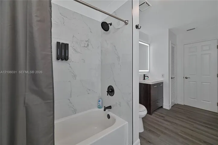 Bathroom featuring a tub with large-format light-toned tile surround, matte black shower fixtures, wood-finish vanity with a rectangular sink, illuminated mirror, and wood-finish flooring