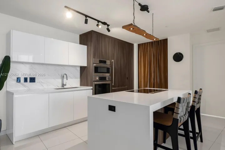 Kitchen featuring white high-gloss flat-panel cabinetry, stone-patterned backsplash, wood-finish floor-to-ceiling cabinetry with built-in stainless steel double wall ovens, center island with white countertop and inset cooktop, and track lighting