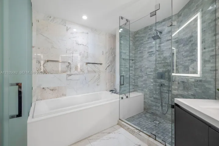 Bathroom featuring a deep soaking tub, walk-in glass-enclosed shower with a built-in bench, textured stone tile wall accents, and an integrated LED-backlit mirror