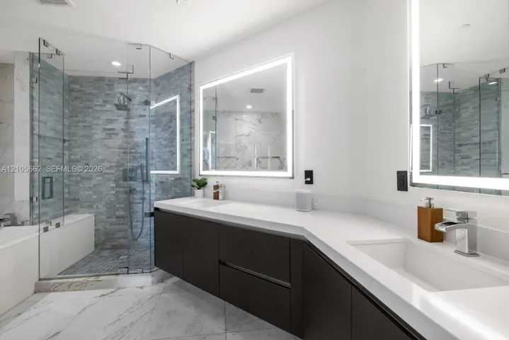 Floating dual-sink vanity with matte dark cabinetry and white countertops, glass-enclosed walk-in shower with textured tile surround, and dual LED-backlit mirrors