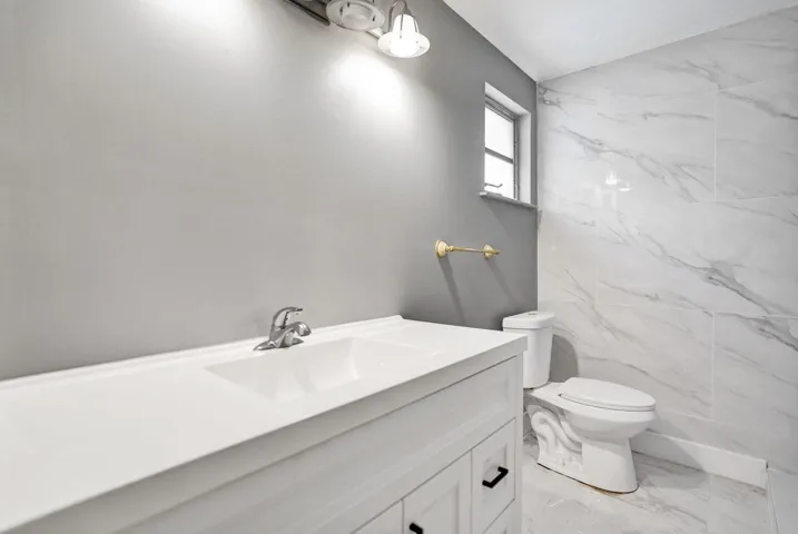 White vanity with a solid-surface countertop and integrated sink, large-format tiled wall surround, two-piece toilet, and multi-light vanity fixture