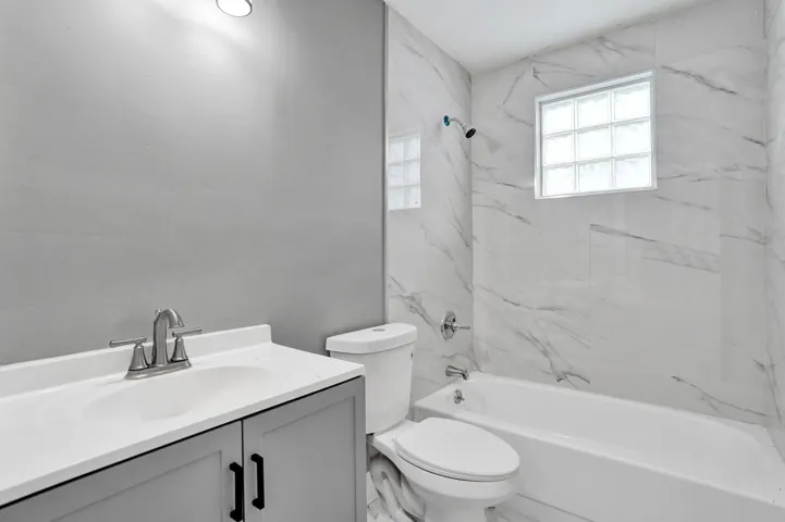 Bathroom featuring a vanity with a solid-surface countertop, gray cabinetry with matte black hardware, bathtub with tile surround, and a glass block window
