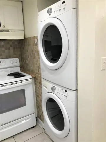 Stacked laundry unit, white electric range with four-burner cooktop, upper cabinetry, and mosaic tile backsplash