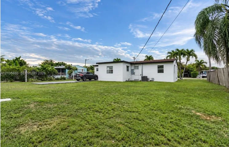 Single-story exterior featuring white stucco siding, a flat roofline, a concrete driveway, and a large open grass yard enclosed by a wood-plank fence