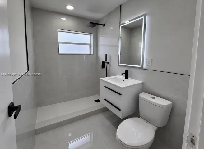 Bathroom featuring large-format gray tile wall and floor surfaces, a floating vanity with a matte black faucet, an LED-backlit mirror, and a walk-in shower with a matte black rainfall showerhead