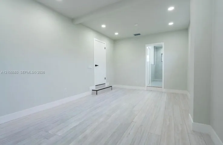 Neutral-toned interior space featuring light-colored wood-finish flooring, recessed ceiling lighting, and a solid-core door with matte black hardware