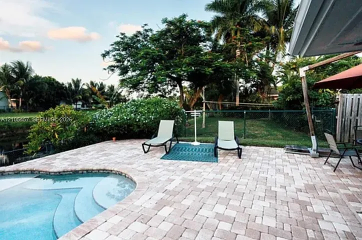 Paver patio featuring a built-in swimming pool with rounded entry steps, perimeter chain-link fencing, and mature palm and shade trees