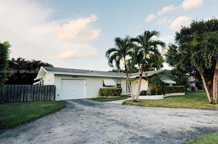 Exterior facade featuring a single-car garage, white stucco siding, metal window awnings, and mature palm trees within the landscaped front lawn