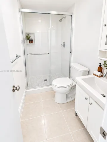 Walk-in shower with sliding glass doors, white tile surround, built-in recessed shelving, vanity cabinet with integrated sink, and large-format tile flooring