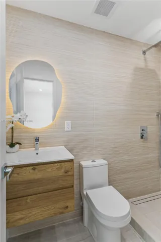 Floating wood-finish vanity with an integrated sink, backlit organic-shaped mirror, wall-mounted shower fixture, and textured stone-finish wall tile