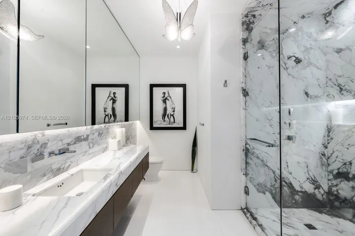 Stone-surfaced vanity with an integrated basin, wall-mounted faucet, expansive wall-to-wall mirror, walk-in shower with glass enclosure, and decorative overhead lighting fixture