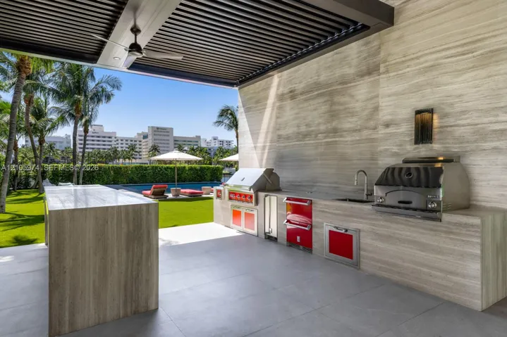 Covered outdoor kitchen featuring stone-clad surfaces, stainless steel built-in grill and beverage refrigeration, integrated faucet, and an overhead louvered pergola with a ceiling fan