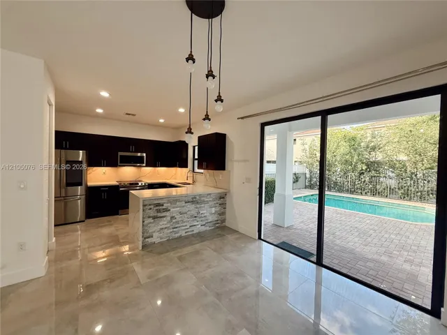 Kitchen featuring dark cabinetry, stone-veneer peninsula, recessed lighting, and large-format tile flooring, accompanied by expansive sliding glass doors opening to a paved patio and swimming pool