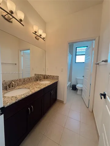 Dual-sink vanity with stone-finish countertop, dark wood-finish cabinetry, full-width vanity mirror, tile flooring, and integrated lighting fixtures