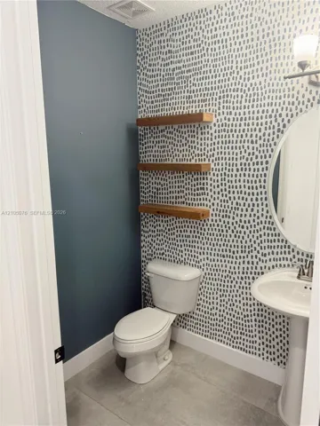 Patterned wallpapered accent wall, three built-in wood-finish floating shelves, white pedestal sink with metallic faucet, oval wall-mounted mirror, and tile flooring