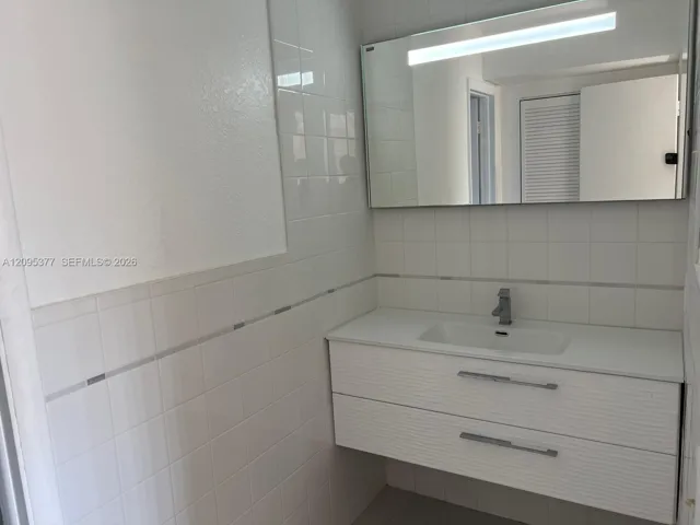 Floating vanity with a white countertop and integrated sink, complemented by a large wall-mounted rectangular mirror featuring an integrated light fixture and white tiled walls