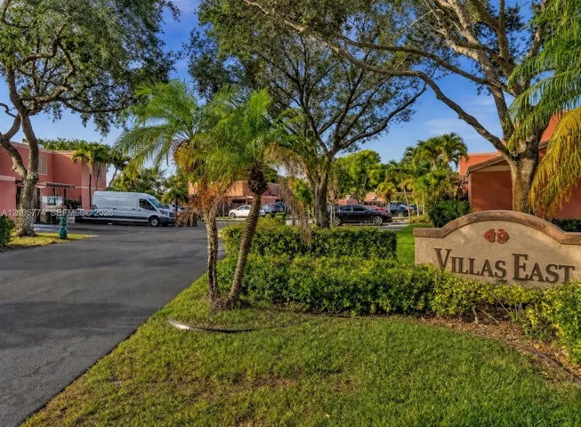 Entryway featuring a monument sign, paved driveway, mature trees, manicured hedges, and palm trees