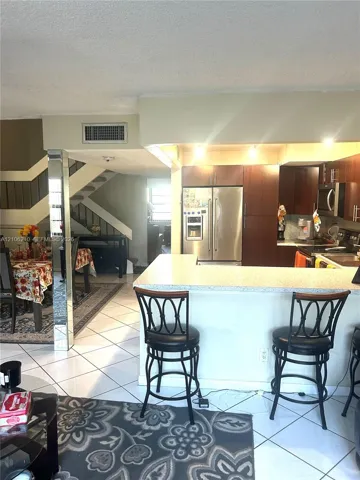 Kitchen featuring wood cabinetry, stainless steel appliances, and a breakfast bar with light-toned stone-finish countertop, accented by polished mirrored support pillars and white tile flooring