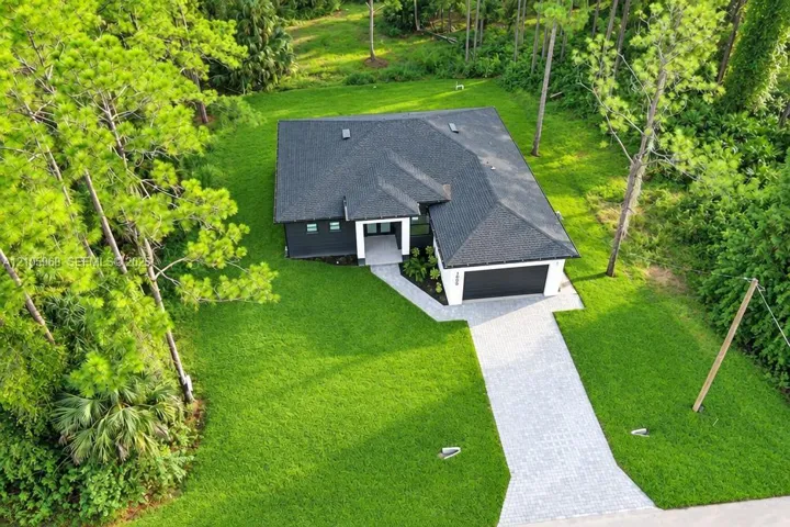 Aerial perspective featuring a single-story residence with a multi-pitched shingled roof, a paved paver driveway, and an expansive lawn bordered by mature trees