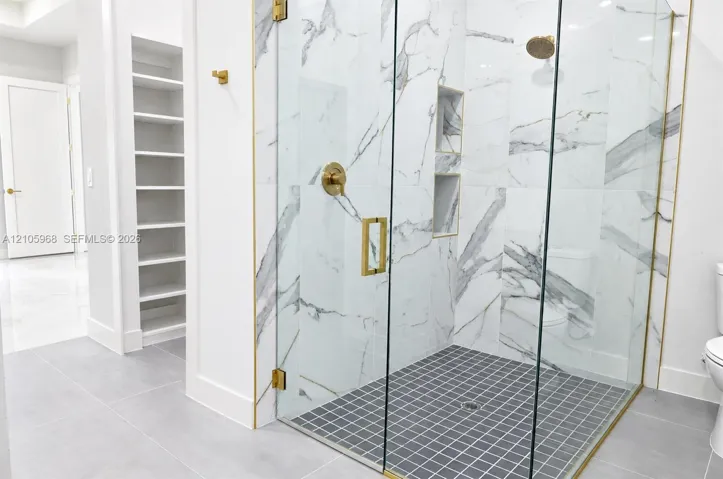 Walk-in shower featuring gold-toned hardware, floor-to-ceiling tiled walls with marbled pattern, recessed wall shelving, and a glass enclosure, complemented by built-in wall cabinetry