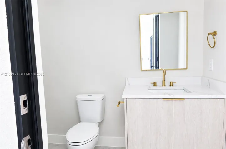 Vanity featuring a light wood-finish cabinet, stone-surface countertop, and gold-toned hardware, accompanied by a rectangular mirror with a gold-toned frame and a gold-toned widespread faucet