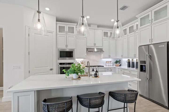 Kitchen featuring white cabinetry with glass-front uppers, solid-surface countertops, a large center island, stainless steel appliances, and three clear glass pendant lights