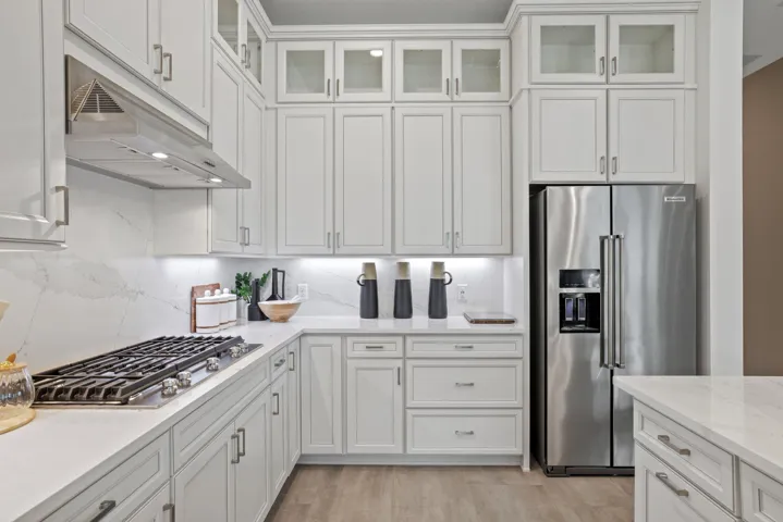 White shaker-style cabinetry with glass-front uppers, light-toned stone-finish countertops, full-height stone-finish backsplash, stainless steel gas cooktop with matching range hood, and integrated stainless steel refrigerator