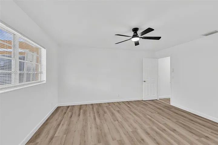Neutral interior featuring wood-finish flooring, white walls, a multi-blade ceiling fan with integrated lighting, and a six-panel interior door