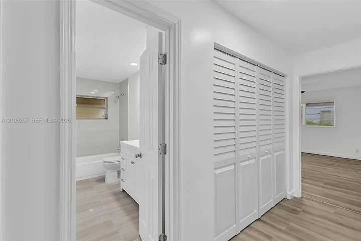 Hallway featuring louvered closet doors, wood-finish flooring, and a bathroom entry with a white vanity and tiled shower enclosure