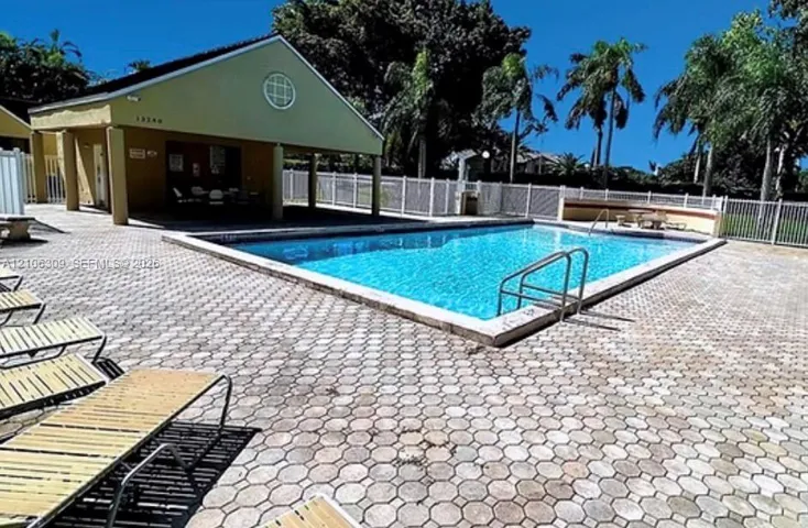 In-ground swimming pool with metal ladder entry, surrounded by a paved deck featuring stone-style pavers, enclosed by a perimeter fence, and accompanied by a covered pavilion structure