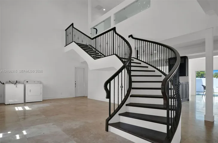 Curved staircase featuring dark wood treads and a black metal railing, complemented by double-height ceilings, stone tile flooring, and a glass-paneled interior balcony