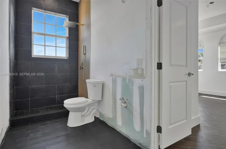 Walk-in shower enclosure with large-format dark tile, wall-mounted rain shower head, dual-pane grid window, and wood-finish flooring