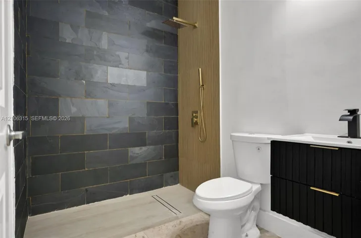 Walk-in shower featuring dark stone-style tile, gold-finish hardware, and a wood-finish panel accent, paired with a black floating vanity and integrated sink