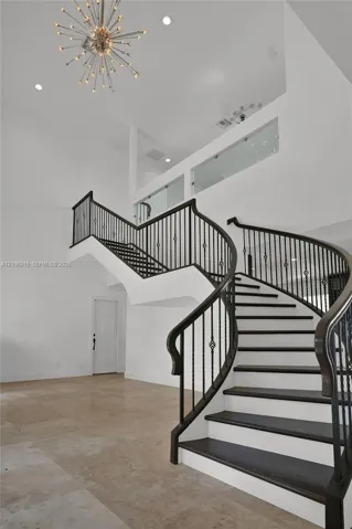 Curved staircase featuring dark-finished treads, white risers, and decorative metal balusters, complemented by high ceilings, a starburst chandelier, and neutral stone-finish flooring